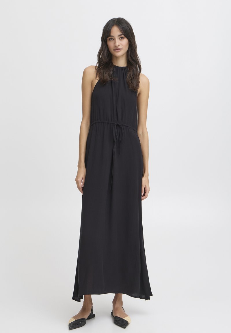 Femme portant une robe maxi noire sans manches avec un col froncé et une ceinture à la taille, associée à des chaussures plates noires et beiges, debout sur un fond blanc.