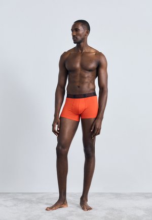 Boxers curtos vermelhos com uma cinta preta exibindo o nome da marca. O tecido parece suave e elástico, ajustando-se de forma justa.