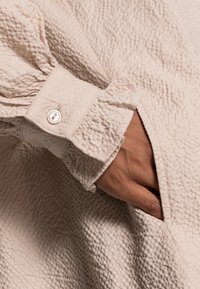 Tejido beige texturizado con un diseño arrugado, que presenta un puño abotonado y una mano en un bolsillo lateral.