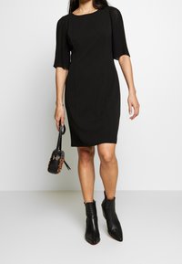 Czarna sukienka do kolan z plisowanymi krótkimi rękawami, okrągłym dekoltem i dopasowanym krojem, w połączeniu z czarnymi botkami za kostkę i małą torebką.
