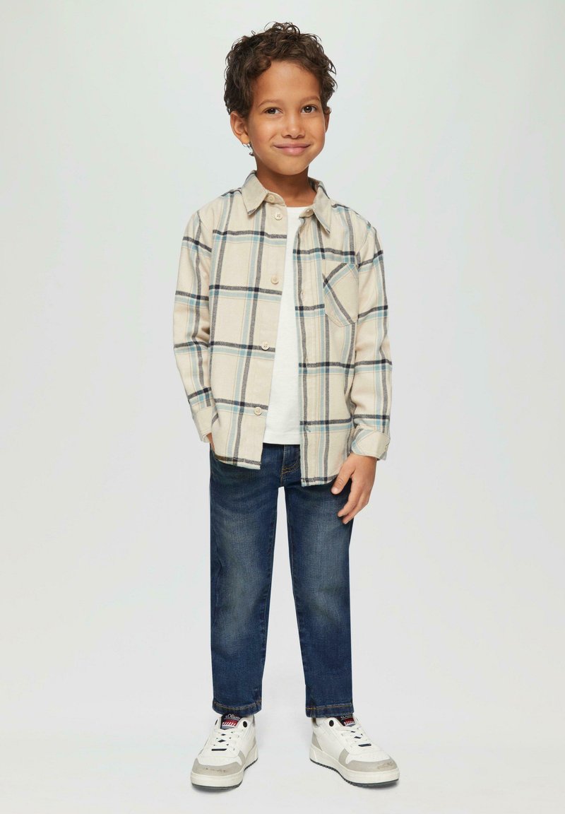 Beżowa koszula w kratę z niebieskimi akcentami, biała koszulka pod nią, ciemnoniebieskie dżinsy i białe trampki. Prosty design i luźny krój.