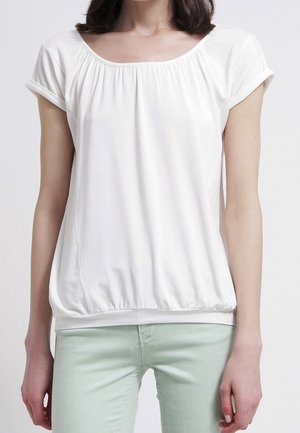 Femme portant un chemisier blanc à manches courtes avec un col et un ourlet froncés, associée à un pantalon vert clair, debout devant un fond uni.