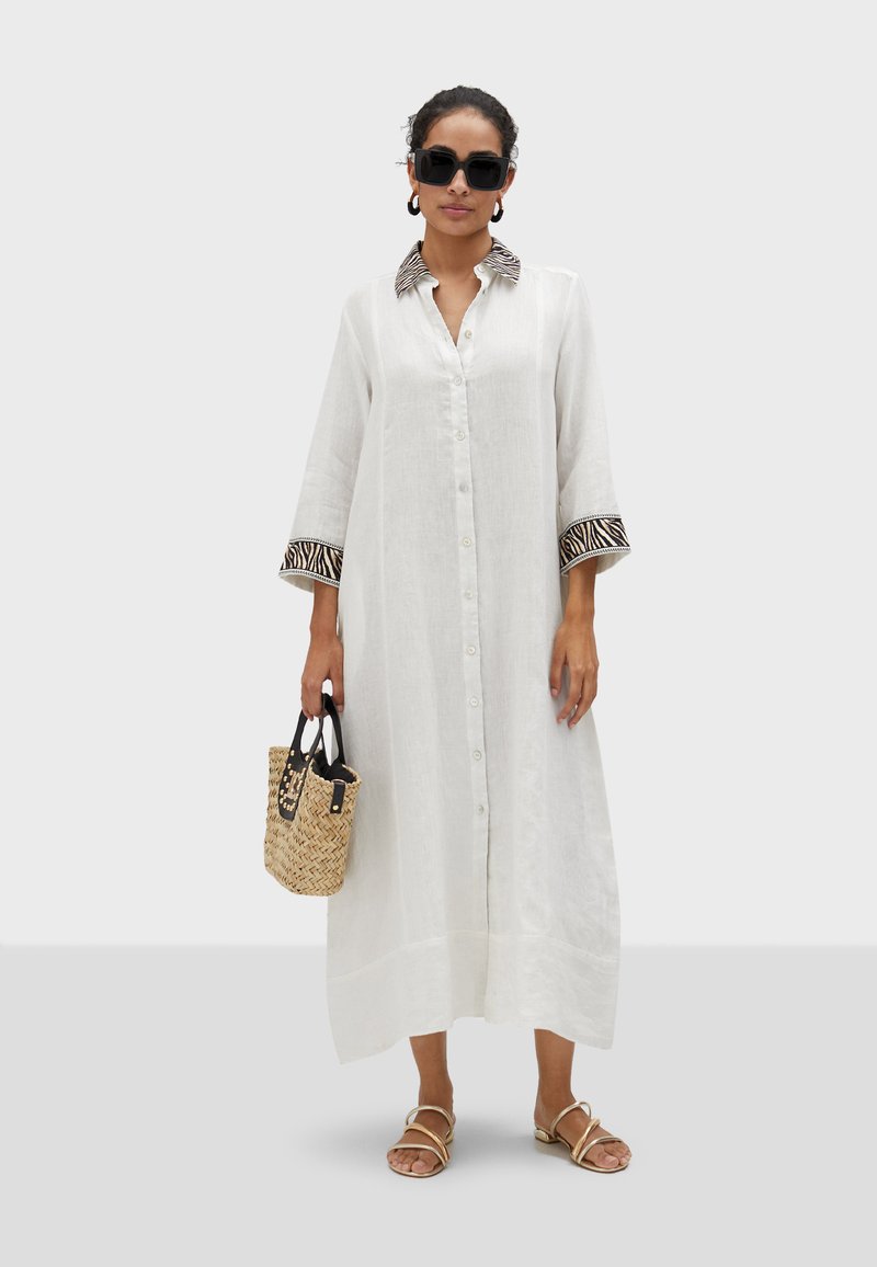 White linen button-up dress with three-quarter sleeves, featuring a zebra-patterned collar and cuffs. Paired with a woven bag and sandals.
