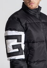 Chaqueta acolchada negra con un cuello alto, que presenta una manga blanca con un gran patrón geométrico en negro. Acabado liso y brillante con cierre de cremallera.