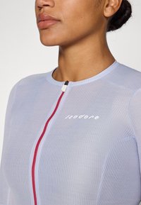 Jasnoniebieska sportowa bluzka z teksturowanej tkaniny, z zamkiem błyskawicznym wykończonym czerwonymi detalami i białym logo "Isadore" na klatce piersiowej.