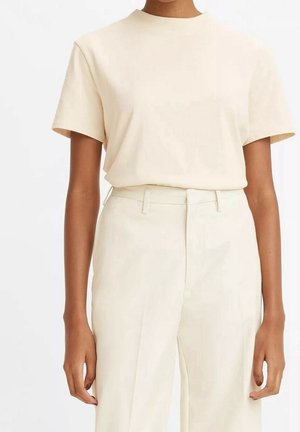 Henkilö pukeutuneena vaalean beigeen lyhythihaiseen, pyöreäkauluksiseen t-paitaan, joka on sullottu korkeavyötäröisiin, kermaan vivahtaviin housuihin, joissa on vyölenkit.