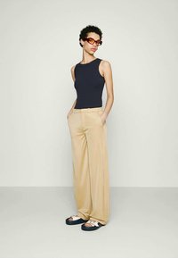Persona che indossa una canotta nera senza maniche, pantaloni beige a gamba larga, sandali bianchi e occhiali da sole con sfumature arancioni, in piedi con le mani nelle tasche.