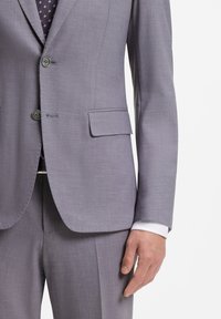 Άνδρας που φοράει ανοιχτό γκρι κοστούμι με σακάκι δύο κουμπιών, τσέπη με καπάκι, μανίκι άσπρου πουκάμισου και γραβάτα με σχέδιο σε λευκό φόντο.