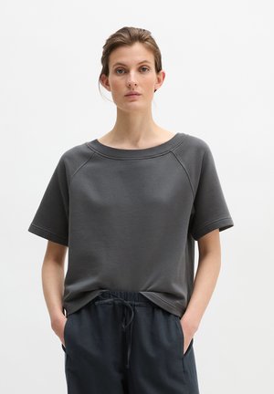 Frau trägt ein dunkelgraues Sweatshirt mit kurzen Ärmeln und marineblaue Hose mit Kordelzug, steht mit den Händen in den Taschen vor einem schlichten weißen Hintergrund.