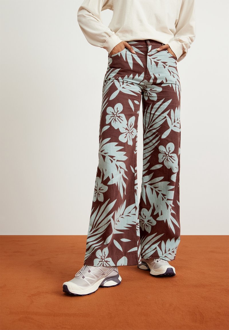 Pantalon à jambes larges en tissu marron orné d'un motif floral turquoise, assorti à des chaussures de sport blanches et violettes.
