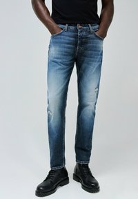 Jeans de mezclilla en un degradado de azul claro a oscuro, con acentos desgastados y un corte recto, combinados con botas negras de cordones.