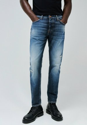 Jeans de mezclilla en un degradado de azul claro a oscuro, con acentos desgastados y un corte recto, combinados con botas negras de cordones.