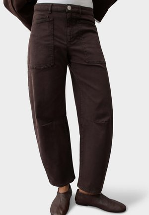 Personne portant un pantalon ample marron foncé avec de grandes poches avant et des chaussures marron foncé à enfiler sur un fond uni.
