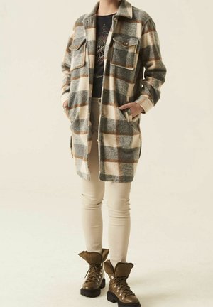 Plaid flannel shirt jacket in cream, grey, and brown, featuring a button-up front, two chest pockets, and side pockets. Styled with beige pants and brown boots.