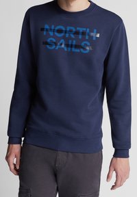 Muž v námořnickém modrém svetru s nápisem "NORTH SAILS" vytištěným tučně modrými písmeny překříženými černými lesklými pruhy.