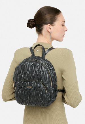 Donna con i capelli raccolti in un bun, che indossa un top a coste beige e uno zaino in pelle nera trapuntata con cerniere dorate e logo sulla schiena.