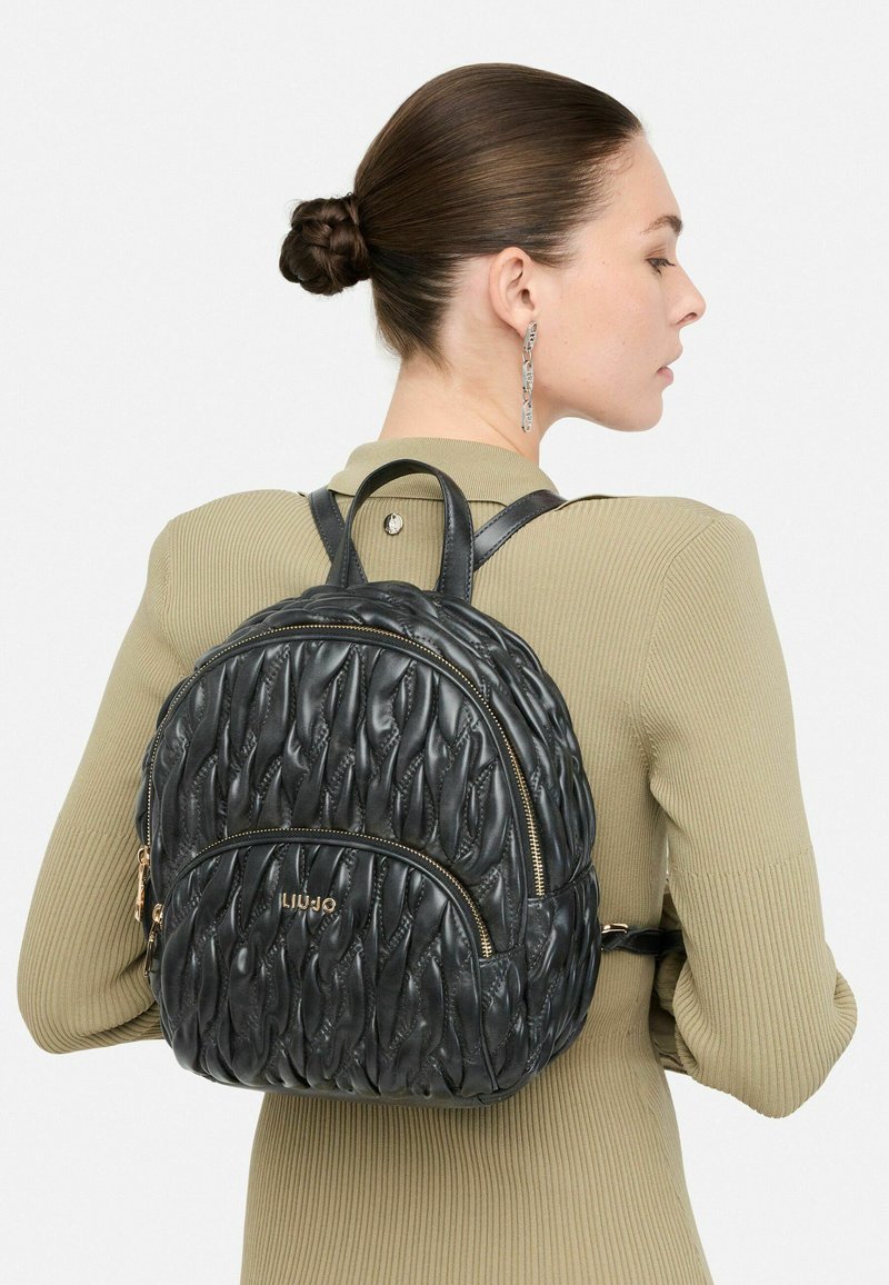 Mujer con el cabello recogido en un moño, vistiendo un top de canalé beige y una mochila de cuero acolchado negro con cremalleras doradas y logo en la espalda.