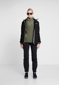 Svart vattentät jacka över en grön fleece-tröja med snoddar, i kombination med svarta byxor och svarta sneakers. Solglasögon som accessoar.