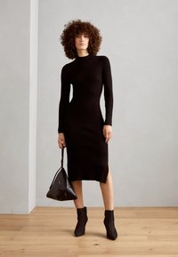 Vestido midi negro acanalado con mangas largas y abertura lateral, combinado con botas negras hasta el tobillo y un bolso triángulo. Textura suave y diseño ajustado.