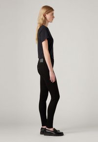 Zwarte aansluitende jeans gecombineerd met een donkerblauwe t-shirt met korte mouwen. Het model draagt zwarte instappers en heeft lang, stijl haar. Minimalistisch ontwerp.