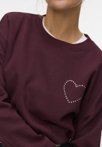 Bordeauxrode trui van zachte stof, met een hartvorm omrand door strassteentjes aan de linkerkant en een geribde halslijn.