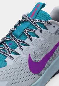 Κοντινό πλάνο λευκού και τυρκουάζ παπουτσιού για τρέξιμο με μωβ λογότυπο Nike και κορδόνια με ασπρόμαυρο σχέδιο. Το κείμενο «Nike Wildhorse» σε κόκκινο χρώμα πάνω στη γλώσσα του παπουτσιού, που είναι τυρκουάζ.