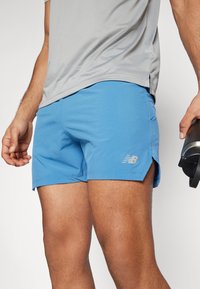 Calções de desporto azuis com textura em rede, fendas laterais e logotipo refletor da New Balance; combinados com uma camisa cinza que elimina a humidade.