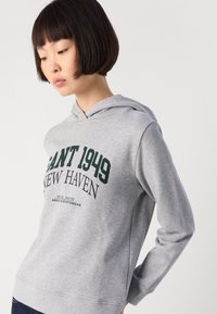 Jeune personne avec les cheveux courts et foncés portant un sweat à capuche gris avec le texte "GANT 1949 NEW HAVEN", regardant vers le bas, se tenant contre un fond uni.