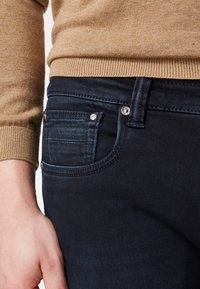 Tmavě modré džíny vyrobené z denimu, s přední kapsou, stříbrnými detaily a hladkým povrchem. Nosit se hodí s světle hnědým pleteným svetrem.