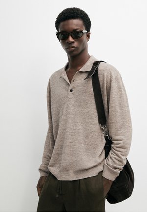 Junger Mann mit dunkler Sonnenbrille, beigem Langarm-Poloshirt, dunkler Hose und schwarzer Schultertasche.