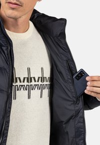 Giacca puffer blu con colletto alto, dotata di una tasca con cerniera. Maglione in maglia avorio sottostante con motivi geometrici neri. Smartphone nero visibile.