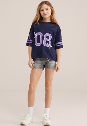 Fată tânără stă în picioare purtând un tricou bleumarin supradimensionat cu "08" și stele, pantaloni scurți gri din denim, șosete albe și adidași albi pe un fundal simplu.