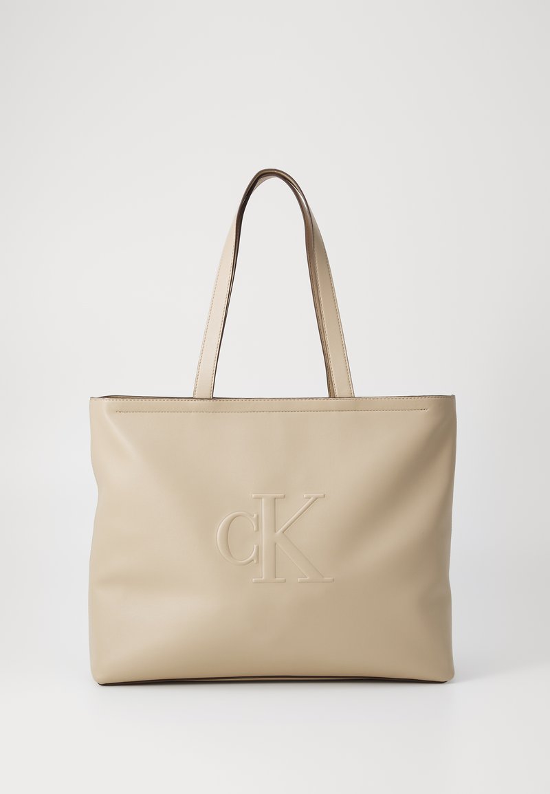 Beżowa skórzana torba typu tote z wytłoczonym logo "CK" z przodu, prostokątna forma, krótkie uchwyty, gładka tekstura, minimalistyczny design.