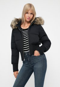 Chaqueta de plumas negra con capucha forrada de piel, diseño corto, puños elásticos y cierre de cremallera, combinada con una camiseta a rayas en blanco y negro y jeans.