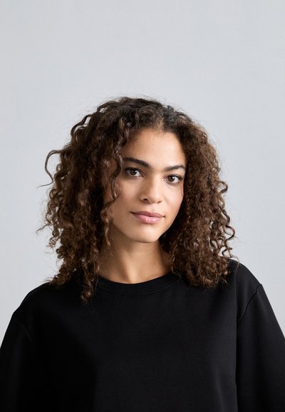 Czarna, oversize bluza z okrągłym dekoltem. Modelka ma kręcone, sięgające ramion włosy. Tło jest jasnoszare, co podkreśla odzież.