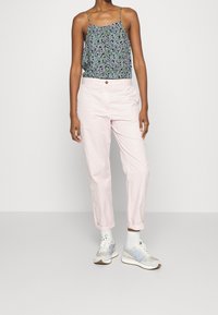 Blomstret top med tynde stropper, parret med lette, lyserøde bukser. Modellen bærer grå og blå sneakers og står foran en neutral baggrund.