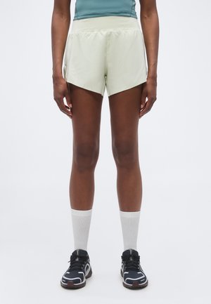 Persoană purtând pantaloni scurți sport de culoare bej deschis, șosete albe până la gleznă și pantofi de alergare negri, stând în fața unui fundal simplu.