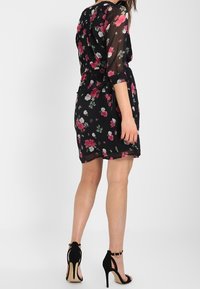 Kvinna i en svart blommig klänning med genomskinliga ärmar, matchad med svarta högklackade sandaletter med ankelsnören, står framför en vit bakgrund.