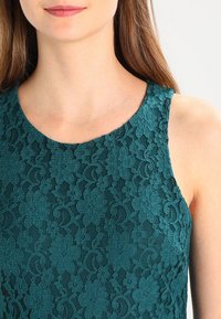 Türkisfarbene Spitze mit rundem Ausschnitt und ärmellosem Design, verziert mit floralen Mustern und einem strukturierten Stoff für einen detaillierten Look.