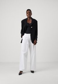 Blazer negro con botón dorado, top negro ajustado, pantalones blancos de pierna ancha y zapatos de tacón alto negros, sobre un fondo claro y liso.