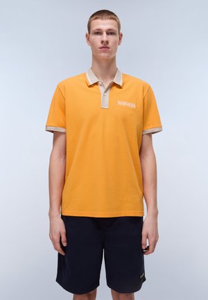 Camisa polo naranja con cuello y puños beige, que presenta un pequeño logo en el pecho, utilizada con pantalones cortos oscuros. Tejido suave y casual.