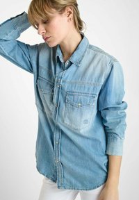 Lichtblauw denimshirt met twee voorszakken, knoopsluiting bij de kraag en lange mouwen. Vervaagt van donkerblauw naar lichtblauw. Zachte textuur.