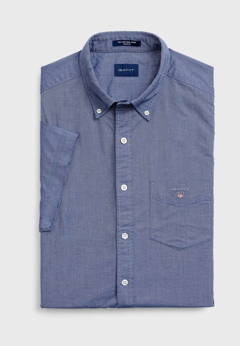 Niebieska koszula z długim rękawem wykonana z bawełny, z kołnierzykiem typu wskazującego, naszywaną kieszenią i małym logo na piersi. Gładka tekstura, regularny krój.