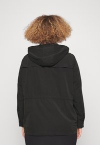 Person med lockigt hår iklädd en svart hooded jacka, sedd bakifrån mot en enkel ljus bakgrund.