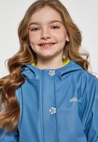 Blå vattentät jacka med huva, med en ljusgrön foder, två tryckknappar och en silverfärgad paraply-logotyp på bröstet.