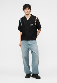 Νεαρός άνδρας στέκεται με τα χέρια πίσω από την πλάτη, φορώντας μαύρο κοντομάνικο πουκάμισο με ριγέ μοτίβα στους ώμους και ανοιχτό γαλάζιο τζιν.