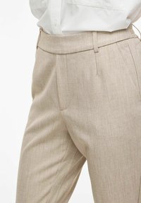 Pantalon beige ajusté avec des plis, porté avec une chemise blanche rentrée, montrant la taille et la partie supérieure de la cuisse.