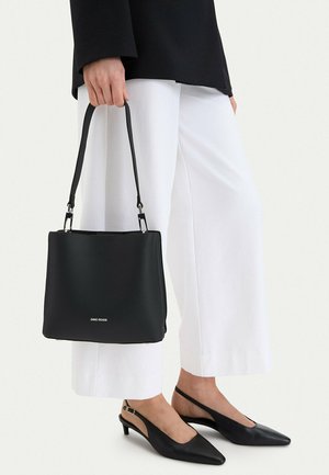 Mujer con tacones slingback negros puntiagudos y pantalones blancos de pierna ancha, sosteniendo un bolso negro de Gino Rossi por el asa.