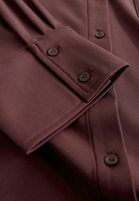 Burgundy long-sleeve shirt with a smooth texture. Features a folded cuff, button details, and subtle pleats on the sleeve.