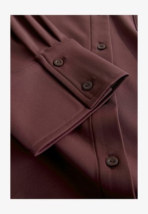 Burgundy long-sleeve shirt with a smooth texture. Features a folded cuff, button details, and subtle pleats on the sleeve.